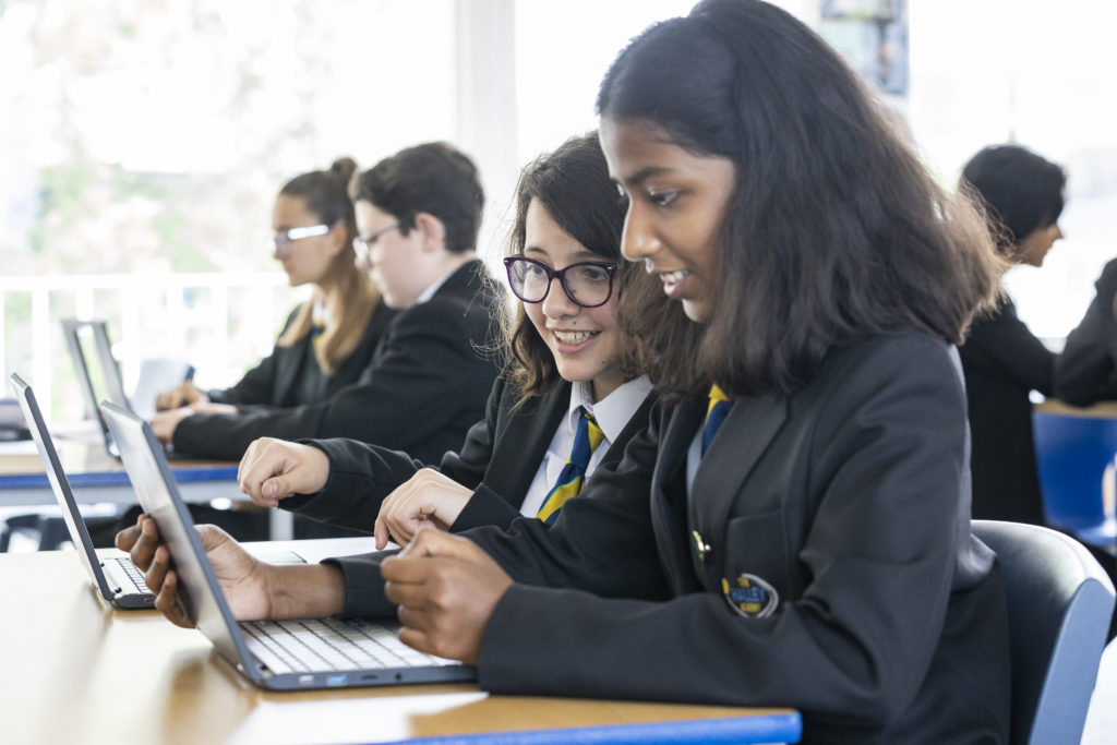 A class of students are shown sitting at their desks and working together, whilst using laptop computers.